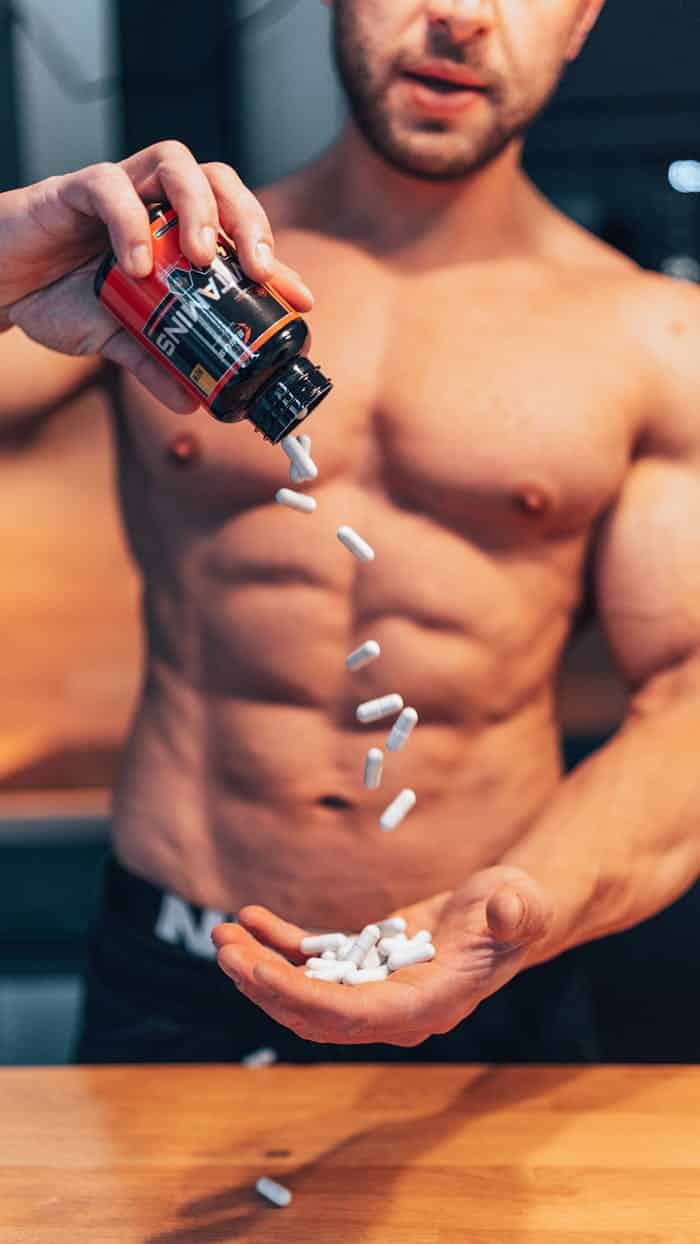 Produtos Shirtless man pouring white supplement capsules into his hand with muscles in focus.