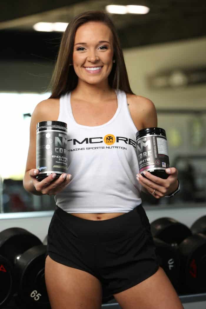 Sobre Nós Smiling woman in gym holding sports nutrition supplements, showcasing active lifestyle.